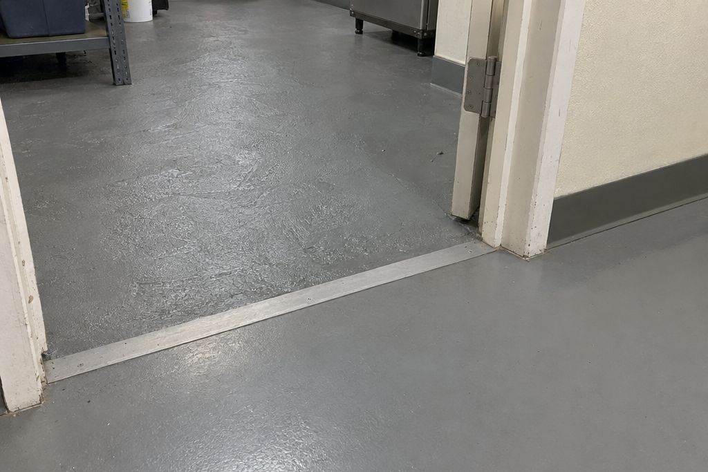 Doorway transition where a thick troweled epoxy mortar floor meets a thinner coated floor at a metal threshold